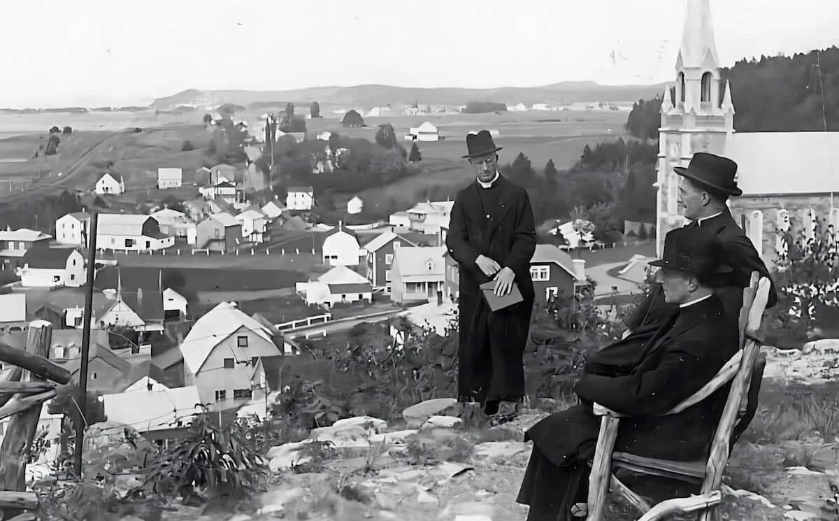 Three Eudist priests in front of the village and church at Pointe-au-Père